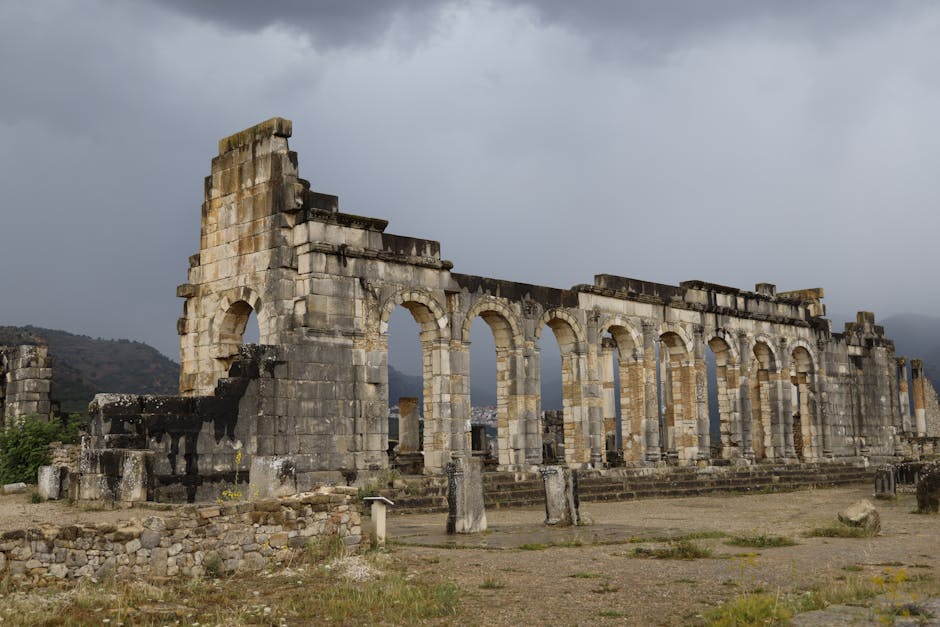 Ancient Roman ruins of Volubilis in Morocco, showcasing stone arches and dramatic cloudy sky.